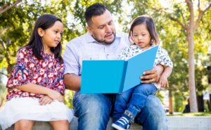 Father sitting on a bench reading a book to his two kids, a daughter and son with down syndrome